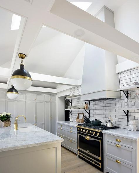 Illuminated by skylights fitted to a sloped ceiling, this gorgeous gray and white kitchen boasts a white hood mounted to white subway backsplash tiles accented with black grout above a La Cornue CornuFe Range and between iron and marble shelves. Range Hood On Sloped Ceiling, Range Hood With Vaulted Ceiling, Patagonia Kitchen, Range Hood Vaulted Ceiling, Kitchen Black And White, Kitchen Cabinets Painted Grey, Black And White Backsplash, Marble Shelves, Grey Painted Kitchen