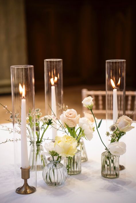 Three taper candles in hurricane covers with pretty flowers in glass bud vases for a wedding table centrepiece. Tapered Candles In Glass Cylinders, Floral Wedding Arrangements, Table Centrepiece Wedding, Wedding Table Decorations Candles, Votive Centerpieces, Christmas Wedding Table, Bud Vases Wedding, Wedding Table Centrepiece, Bud Vases Arrangements