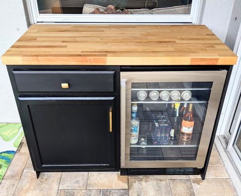 Outdoor bar with wooden countertop, black cabinet and drawer with gold handles on the left and stainless steel glass front mini-fridge on the right Deck Fridge Ideas, Mini Fridge Outdoor Patio, Outdoor Bar With Mini Fridge, Mini Fridge Cabinet Beverage Center, Porch Mini Bar, Outdoor Bar Fridge Ideas, Glass Front Mini Fridge, Diy Outdoor Bar With Mini Fridge, Diy Wine Cabinet With Fridge