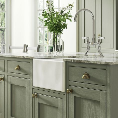 A muted green kitchen with brass handles a white farmhouse sink with chrome sink and solid surface worktop with flowers in a vase Symphony Kitchen, Brass Kitchen Accessories, Green Shaker Kitchen, England Kitchen, Light Green Kitchen, New England Kitchen, Brass Kitchen Handles, Open Plan Kitchen Dining Living, Open Plan Kitchen Dining