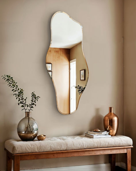 The image showcases the Abstract Wave Wall Mirror, a modern mirror with an intriguing irregular shape that adds a touch of artistic flair to the room. The mirror is mounted above a wooden bench with a soft, cushioned top, adorned with decorative items including a large glass vase with greenery, a metallic vase, and a small stack of books. The overall decor creates a warm and inviting ambiance, blending contemporary design with a cozy, natural feel. Irregular Shape Mirror, Shape Mirror, Mirror For Living Room, Unique Mirror, Wavy Mirror, Wave Wall, Hallway Inspiration, Unique Mirrors, Abstract Waves