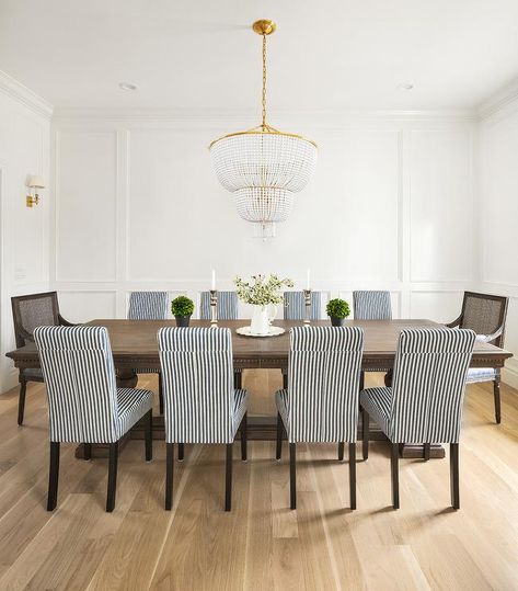 Black and white stripe dining chairs and cane back captain's chairs surround a dark stained French dining table illuminated by an Aerin Jacqueline 2-Tier Chandelier, while wainscot walls are lit by brass sconces. Klismos Dining Chair, The Fox Group, Striped Dining Chairs, Oval Wood Dining Table, French Dining Table, Fox Group, Light Wood Dining Table, Bamboo Dining Chairs, French Dining Tables