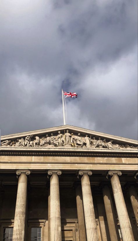 Zommed picture of the top entrance of the British Museum. The UK flag is waving to the wing. It’s a cloudy dark day London Flag, British Aesthetic, Aesthetic London, Museum Aesthetic, London Aesthetic, Uk Flag, Dark Clouds, British Flag, England Uk