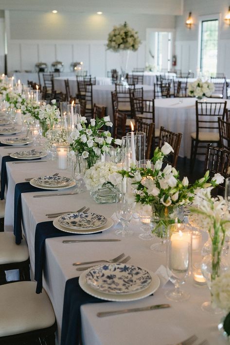 Elegant Wedding Reception Decoration | A refined and elegant reception table setup featuring white tablecloths with navy blue table runners. The table is adorned with blue and white patterned plates, sleek silver flatware, and crystal glassware. Complemented by lush floral arrangements of white roses, hydrangeas, and greenery in clear vases. Dark wooden chairs add a sophisticated contrast, and the white linens and stunning tall floral centerpieces make for a cohesive, timeless design. Navy Table Setting Wedding, Blue Wedding Theme Fall, Blue Table Settings Wedding, Tall Floral Centerpieces, Blue Table Runners, Navy Blue Table Runner, Navy Wedding Colors, Blue And White Wedding, White Linens