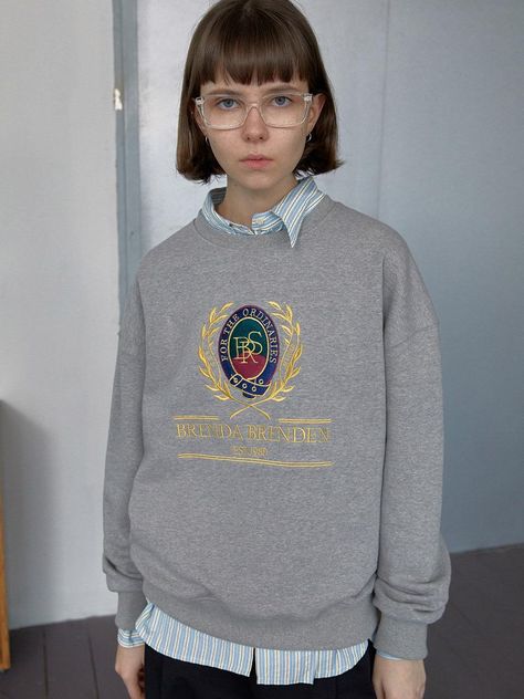 The Classic Sweatshirt depicted here offers a blend of traditional collegiate style and modern casual wear. It features a distinctive crest graphic on the front, with elaborate details and text that reads BRENDA BRENDEN EST. 1980, adding a touch of preppy flair. The gray hue of the sweatshirt makes it a versatile piece that can be easily paired with various outfits, while its crew neck and ribbed cuffs ensure a comfortable and snug fit. - The crest graphic is richly designed, making the sweatshirt stand out as a statement piece while still retaining a classic look.- Made from soft, durable fabric, this sweatshirt provides warmth and comfort, ideal for cooler days.- Its traditional fit and simple color allow it to be styled effortlessly with both casual jeans and more formal trousers.- Collegiate Sweatshirt Outfit, Sweat Shirt Outfits Women, Graphic Crewneck Outfit, Collared Shirt Under Sweatshirt Outfit, Shirt Under Sweatshirt, Grey Sweatshirt Outfit, College Sweatshirt Outfit, Crew Neck Sweatshirt Outfit, Gray Sweatshirt Outfit