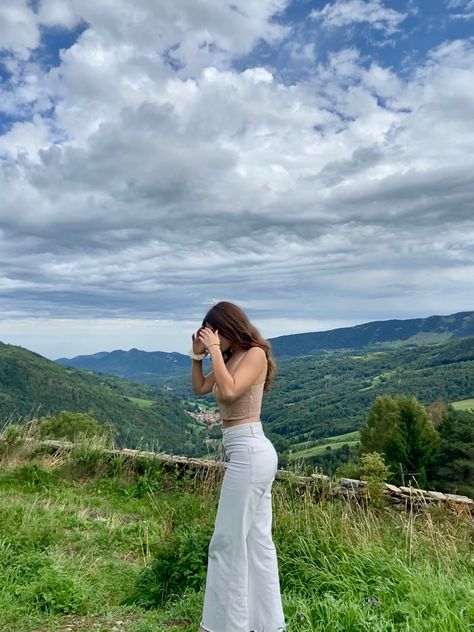 Young girl in nature looking down with hands on face. Mountains in the backgrownd. Brown long hair, beige corset top, lilac cardigan and white flared pants. All green nature and cludy, moody sky. Mountain Outfit Aesthetic, Aesthetic Photos Nature, Hill Station Outfit Ideas, Mountain Photography People, Mountain Outfit Summer, Mountain Aesthetic Outfit, Mountain Photo Ideas Instagram, Corset Top Outfit Ideas, Mountain Poses Photo Ideas