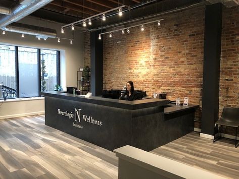 This black desk really stands out against the light wooden floor. Exposed brick long with the open ceiling and track lights adds an industrial vibe. #design #chiropractic #lobby Brick Interior Office, Office Exposed Brick, Industrial Chic Lobby, Exposed Brick Office Space, Front Desk Office Design, Office Wooden Flooring, Industrial Clinic Design, Brick Office Design, Lobby Industrial Design