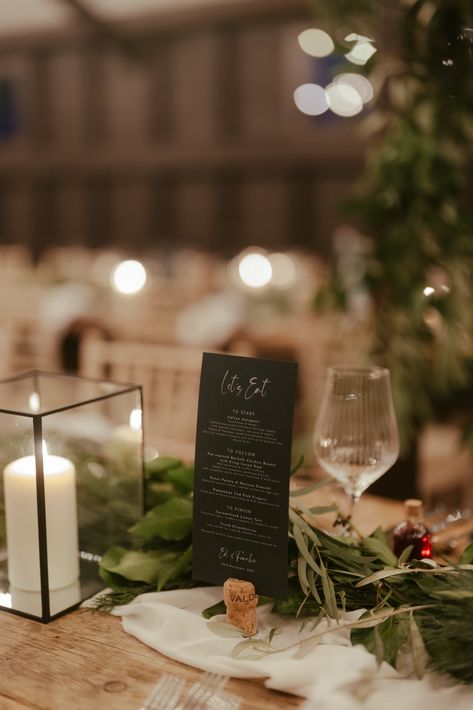 This shows a close up of a dining set up at a wedding with modern rustic vibes. On the table is a black box lantern with a glowing pillar candle in it. Next to it is a black simple stationary showing the menu. There is also a white table runner, greenery and glassware. Black Stationary, White Table Runners, Wedding Dining Table, Glass Lanterns, Wedding Dining, Barn Photography, Wedding Lanterns, Glass Lantern, White Table