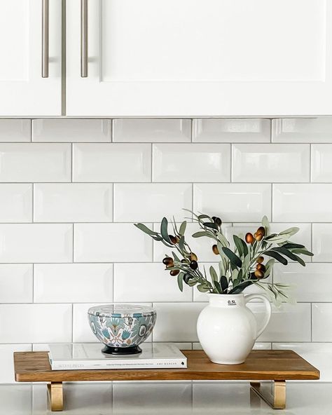 A white kitchen cabinet hangs over a white subway tile backsplash with light grey grout. Green branches in a white pitcher sit on a wooden tray next to a blue bowl on a white book. The tray is placed on the white countertop. Kitchen Counter Styling Ideas, Kitchen Wall Covering, Counter Styling, White Kitchen Counters, Kitchen Counter Styling, Modern Kitchen Counters, Gray Grout, Warm Wood Flooring, Light Gray Cabinets