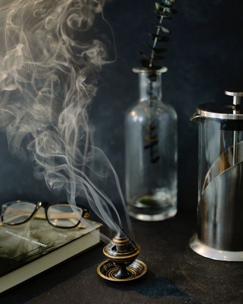 Smoke coming out of a black incense burner with gold details against a black background. You can also see a book with glasses on top, a glass bottle with eucalyptus and a French press Goth Product Photography, Dark Still Life Photography, Gothic Product Photography, Spooky Product Photography, Witchy Product Photography, Dark Background Photography, Halloween Product Photography, Moody Product Photography, Aesthetic Incense