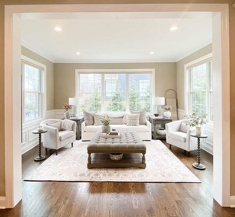 Stroll into this spacious living room with beige walls and white wainscoting. A decorative rug hosts a tufted ottoman. Light gray armchairs and a white sofa are gathered around the ottoman, creating an ideal environment to chat with your family or guests. Living Room With Beige Walls, Sunroom Interior, Room With Beige Walls, White Wainscoting, Room Wall Colors, White Sofa, Beige Wall, Grey Armchair, Tufted Ottoman