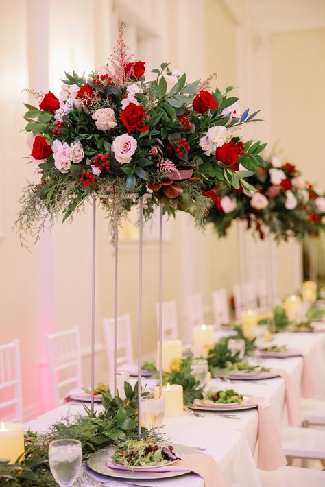 Long table with white table cloth, plated salad and blush napkin. Lush winter greenery runner lines the center of the tablescape, with tall abundant centerpieces throughout. Red Floral Table Centerpieces, Red Rose Centrepiece Wedding, Tall Red Flower Arrangements, Pink Red White Flower Centerpiece, Red And Pink Ceremony Flowers, Winter Wonderland Wedding Centerpieces, Bride Groom Table, Red Wedding Decorations, Winter Wedding Centerpieces