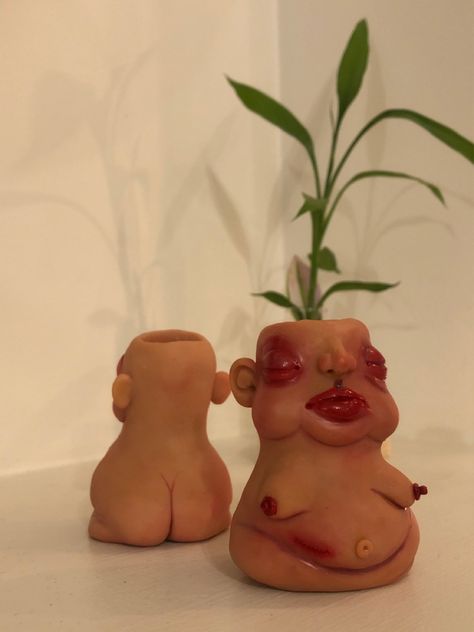 In the foreground, a clay blobby dude with olive tan skin, maroon eyeshadow and puffy red under eyes. Voluptuous red lips, and a scar on the lower stomach simulating a recently removed appendix. Saggy exposed boobs, and pudgy stomach. A tall leafy plant appears to be coming from its head. In the background, a figure of similar height and skin tone, facing away from the camera to show its buttcrack ~ simulating a mirror image.  

Plain white surroundings and warm light. Lighter Holder Clay, Clay Lighter Holder, Clay Lighter, Clay Diys, Lighter Holder, Candle Pot, Clay Candle, Light Clay, Clay Diy Projects