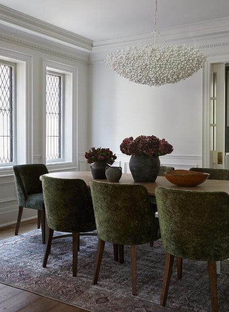 Elegant dining space showcases an Oly Studio Berrie Bowl chandelier hung above an oval brown wooden dining table surrounded by forest green velvet dining chairs placed on a pink and purple rug. Dark Green Chairs Dining Rooms, Brown Velvet Dining Chair, Green Velvet Dining Chairs, Bowl Chandelier, Condo Decor, Transitional Dining Room, Green Dining Chairs, Wooden Dining Table, Condo Decorating