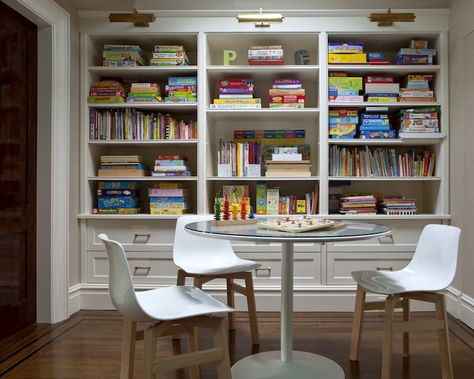 Fantastic game room features wall of built-in bookshelves filled with board games and books illuminated by antique brass picture lights situated behind glass-top game table surrounded by white plastic chairs White Plastic Chairs, Board Game Room, Garage Game Rooms, Homework Room, Basement Games, Board Game Storage, Game Room Basement, Game Room Design, Upper East Side