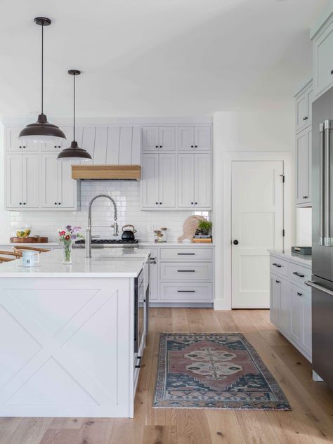 This modern farmhouse kitchen features white tones, natural woods, and stainless appliances, which lend the room a simple, clean aesthetic. Shaker-style cabinetry is adorned with matte black hardware, which is echoed in the matte black pendant lights over the marble topped island. White subway tiles give the space behind the stove a glossy sheen, while a vintage runner adds warmth underfoot. Kitchens Black Hardware, Maine Interiors, Matte Black Pendant Lights, Maine Farmhouse, Maine Kitchen, Black Pendant Lights, White Subway Tiles, Matte Black Hardware, Clean Aesthetic