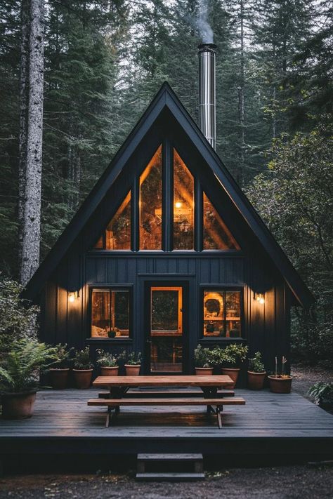 Front view of a modern a frame cabin in charcoal black siding full wall glass windows natural wood door with glass panels tall chromed chimney in. Check out all of these stunning and chic dark A-frame houses that pack a ton of charm into a small forest retreat. A Frame Black House, Moody Modern Cabin, Small Black Cabin, Log Cabin A Frame, A Frame House Ideas, Moody A Frame, Dark Cabin, Black A Frame Cabin Exterior, Glass Cabin In Woods