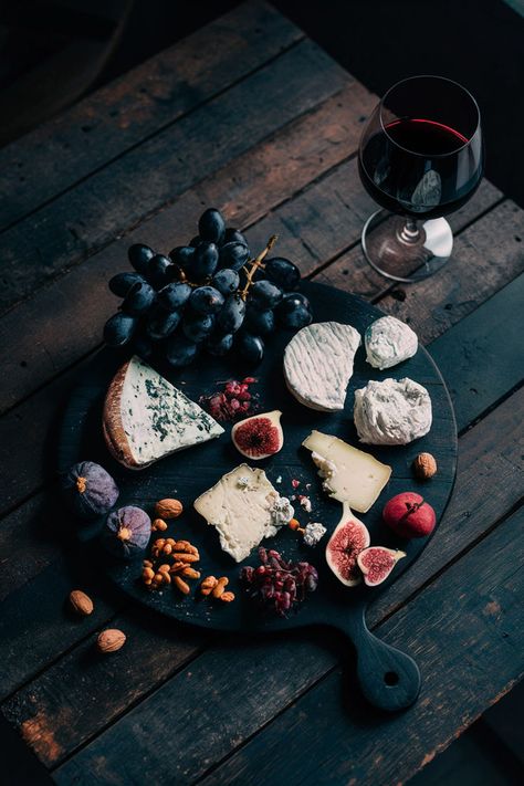 Dark wooden cheese board with assorted cheeses, figs, black grapes, nuts, and a glass of red wine, set on a rustic wooden table with moody lighting. Wine And Cheese Board Aesthetic, Moody Charcuterie Board, Dark Charcuterie Board, Charcuterie Board Photoshoot, Cheese Platter Aesthetic, Wine And Cheese Aesthetic, Dinner Cheese Board, Cheeseboard Aesthetic, Black Charcuterie Board