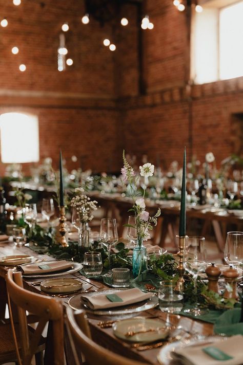 This shows a dining set up, woodlands inspired. The focus is on one of the rows of the wooden dining tables. On it are dinnerware, gold cutlery, a forest green table runner, brass candlesticks and flower vases. Brass Candlesticks Wedding Centerpieces, Taper Candle Decor, Brass Candlesticks Wedding, Candlestick Centerpiece Wedding, Candlestick Collection, Barn Photography, Candle Sticks Wedding, Vintage Brass Candlesticks, Brass Candlestick