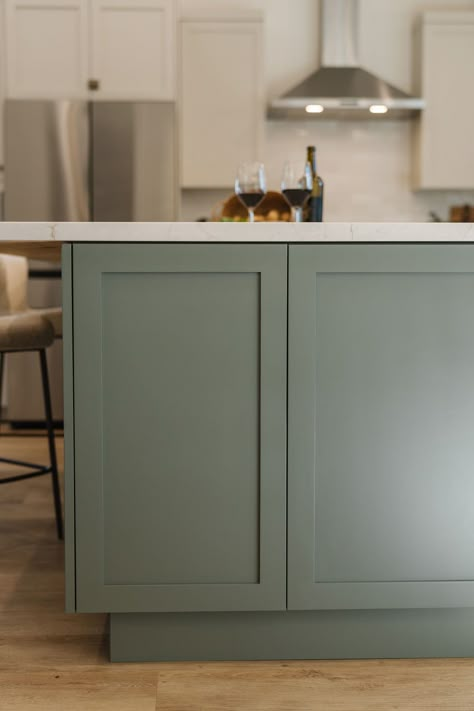 We used Benjamin Moore's Intrigue on this kitchen island, which is a moody gray with green undertones and it adds just a bit of color to this warm toned neutral kithcen. Hidden cabinet in island. #thedendesigngroup #benjaminmoore #moodypaint #kitcheninspo #kitchenisland #whitoakfloors Green Gray Cabinets, Gray Green Cabinet Colors, Green Cabinets Gray Floor, Green Gray Island Kitchen, Moss Green Island Kitchen, Pewter Green Kitchen Island, Green Island Wood Cabinets, Kitchen Cabinet Color Ideas Two Tone Island, Avon Green Benjamin Moore Cabinets