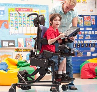 In a classroom setting, a young boy with cerebral palsy is seated in an adaptive chair while his teacher holds up a game for him to play with Activity Chair, Rare Genetic Disorders, Individual Education Plan, Spy Gear, Adaptive Equipment, Accessible Bathroom, Activities Of Daily Living, Wheel Chair, Education Technology