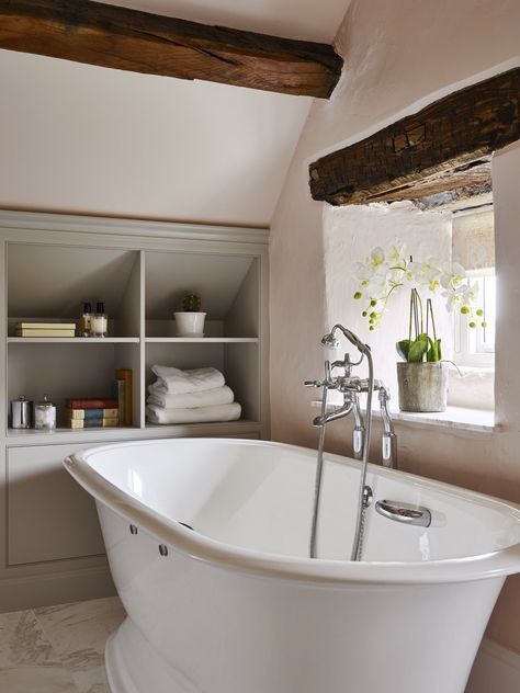 Sloping ceilings and heavy beams can present design challenges in older properties. The bathroom featured was designed to maximise storage with cabinetry and shelving handmade to fit the available space under the eaves. Freestanding bath from @vandabaths, taps and accessories from @samuel_heath and furniture finished in #purbeckstone from @farrowball.⁠ ⁠ #bathrooms #ensuite ⁠ #bathroom #bathroomdesign ⁠ #bathroomcabinetry #bath ⁠ #bathroomdesigners ⁠ #bespokebathroom⁠ #yorkshirebathroom⁠ Bathroom In The Eaves, Eaves Bathroom Storage, Bathroom In An Attic, Bathroom Eaves Storage, Bathroom With Eaves, Bathroom Sloping Ceiling, Bathrooms With Sloping Ceilings, Exposed Beams Bathroom, Shower Under Sloped Ceiling