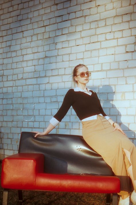 Woman wearing sunglasses, a black sweater over a white button-up shirt and a tan skirt sitting on the back of a red and black mid-century couch. A white brick wall is in the background. Cinestill 400d Portrait, Cinestill 400d, Cinestill 800t, Cinematic Photography, Film Photography, Short Film, Portrait Photography, Fashion Photography, Film