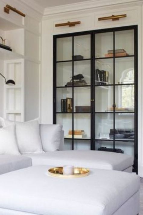 We greatly appreciate a beautiful built-in featuring glass doors! We also love the look of these black-frame glass doors and their contrast to this bright, white living area. Read our blog post for more built-in design inspiration. #interiordesign #interiordesigner #built-ins #built-in #shelves #glassdoors #whitesofa #sofa #shelfstyling #gallerylights #bookshelves Glass Shelves In Bathroom, Glass Shelves Decor, Glass Cabinet Doors, Hus Inspiration, Glass Cabinet, A Living Room, Book Shelf, Cheap Home Decor, Built Ins