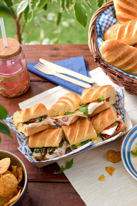 A table outside topped with sandwiches, drinks, cutlery, chips and bread. Sandwich Picnic, Savory Halloween Food, Caramel Waffles, Picnic Photography, Sliced Salami, Sub Rolls, Brioche Rolls, Chilli Jam, Green Pesto