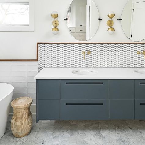 White penny tiles accented with black grout is lined with wood border tiles and finished with antique brass faucets positioned over round sinks fitted to a white quartz countertop complementing a dark gray floating washstand adorned with oil rubbed bronze pulls. Penny Tile Dark Grout Bathroom, Navy Penny Tile, Blue Penny Round Tile Bathroom, Dark Grey Penny Tile Bathroom, Grey Penny Tile, Vanity Backsplash, Minimal Bathroom, Mosaic Bathroom Tile, Modern Backsplash