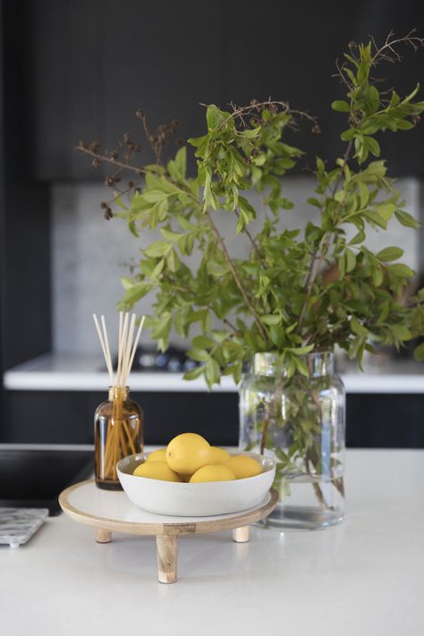 Vignettes or styled arrangements add interest to surface areas of your home. They are also a great way to display your favourite items! You can put them on your coffee tables, sideboards, kitchen benches, beside table, shelves and so much more! We give you the best tips and tricks on how to create and style a vignette in your space. Kitchen vigentte styling with stylised tray, aroma diffuser, bowl of fruits, lemons, large vase, foraged branches. Click the pin to see more vignette inspo and tips! Kitchen Bench Fruit Bowl, Fruit Bowl Decor Centerpieces, Fruit Bowl On Island, Farmhouse Fruit Bowl, Bowl Of Lemons Decor, Lemon Bowl Decor, Fruit Bowl Kitchen Display, How To Display Fruit On Counter, Wooden Risers Display Kitchen