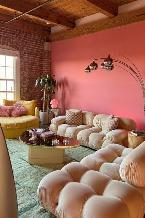 A vibrant living room with a bold pink accent wall, exposed brick, and wooden ceiling beams. It features plush beige tufted sofas, a yellow couch with a pink fluffy pillow, a stylish coffee table with books, and a sleek floor lamp with globe lights. A green plant adds a touch of nature. The room is bathed in natural light from a large window, creating a cozy and chic atmosphere perfect for summer lounging. Loft Apartment Living Room, Pink Accent Wall, Living Room Paint Colors, Patchwork Decor, Pink Accent Walls, Pink Apartment, Girl Apartment, Summer Living Room, Living Room Transformation