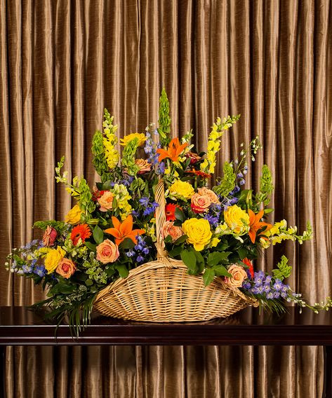 A bold and colorful collection of flowers designed in a fireside basket to celebrate the fond memories of a loved one. Thanksgiving Arrangements, Orange Lilies, Orange Gerbera, Bells Of Ireland, Flower Farming, Altar Flowers, Large Flower Arrangements, Blue Delphinium, Roses Blue