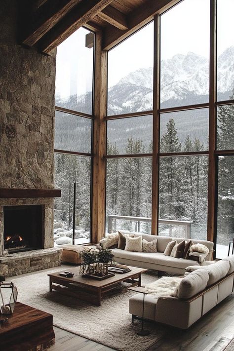 Mountain home living room with exposed rustic beams full glass walls a stone chimney modern furniture winter pine forest and mountain view through the windows. Check out all of these amazing modern rustic mountain houses to blend natural charm with contemporary flair. House With A Mountain View, Mountain Inspired Living Room, Mountain View Window, Home With Mountain View, Mountain View Living Room, Modern Chalet Living Room, Rustic Stone Fireplace Ideas, Montana Cabin Aesthetic, Mountain Cabin Modern