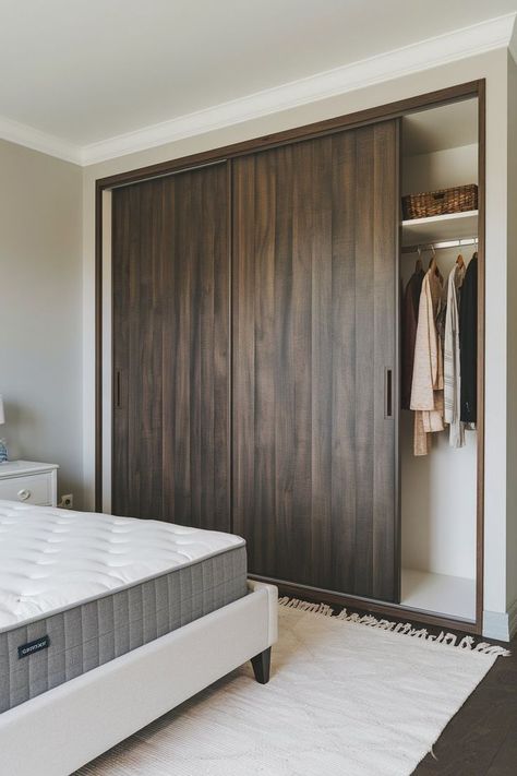 Bedroom with a dark wood sliding door wardrobe, white-framed bed with gray mattress, light gray walls, white rug, and a few clothes hanging in the wardrobe. Wardrobe Dark Wood, Bedroom Design With Wardrobe, Dark Wood Wardrobe, Framed Bed, Wood Sliding Door, Sleek Wardrobe, Sliding Door Wardrobe, Stylish Bedroom Design, Wood Wardrobe