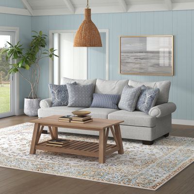 This sofa features a laid-back silhouette with roll arms and pillow backs to bring a hint of coastal farmhouse charm to your living room. It has a solid and engineered wood frame that sits on tapered legs with a dark brown finish. Wrapped with 100% polyester in a neutral gray tone, this couch is filled with foam and polyester and supported by sinuous springs to give you some padding as you relax. The seat and back covers are removable to make cleaning a cinch. Five vibrantly patterned toss pillo Grey Coastal Living Rooms, Coastal Casual Living Room, Coastal Farmhouse Living Room, Beach House Decor Living Room, Hamptons Living Room, Coastal Sofa, Florida Living Room, Beachy Living Room, Coastal Living Room Furniture