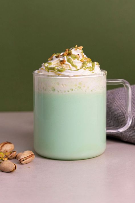 A green coloured pistachio and rose flavoured hot chocolate, made with white chocolate powder. The drink is in a transparent mug, sat on a grey table next to some pistachios, all against a dark green background. The drink is topped with whipped cream, pistachio flavoured sauce and crushed pistachios. Pistachio Hot Chocolate, Romantic Drinks, Pistachio Syrup, White Hot Chocolate Recipe, Chocolate Coffee Beans, Milkshake Machine, Coffee Syrups, Opening A Coffee Shop, Slushie Recipe