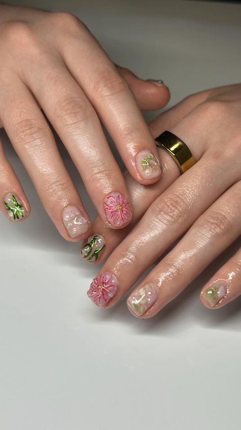 The image shows a close-up of a person's hands displaying an intricate gel nail art design. The nails feature a mix of pink and green botanical themes. Each nail has a unique design, including 3D pink flowers, green leaves, and small pearls on a clear or light-colored base. The person's fingers are well-manicured and appear moisturized, with the nails having a glossy finish. The person wears a large gold ring on the index finger of their left hand. The background is a plain white surface. Fairy Nail Art Short, Pink And Green Short Nails, Pink And Green Nails Short, Pixi Nails, Fairy Nails Short, Short Summer Gel Nails, Funky Short Nails, Nails Memorial Day, Early Summer Nails