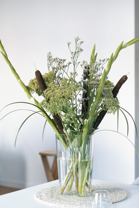 Floral arrangement with broadside cattail, green foliage and pale flowers, arranged in a transparent glass vase on a decorative off-white tablecloth, with a white background Cattail Bouquet, Typha Latifolia, Greenery Floral Arrangement, Pale Flowers, Selling Flowers, Unique Floral Arrangements, Transparent Flowers, Summer Lake, Community Garden