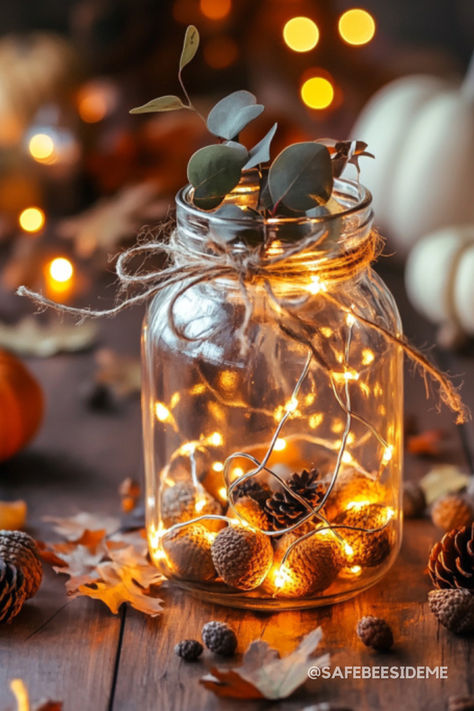 A mason jar centerpiece with warm fairy lights, acorns, pinecones, and eucalyptus sprigs tied with rustic twine, surrounded by autumn leaves and warm candlelight on a wooden table. *We may earn a small commission from your purchase. Mason Jar Fairy Lights Centerpiece, Mason Jar Fairy Lights, Fairy Lights In A Jar, Mason Jar String Lights, Indoor Photoshoot, Gallon Glass Jars, Mason Jar Centerpiece, Fall Mason Jars, Twine Bow