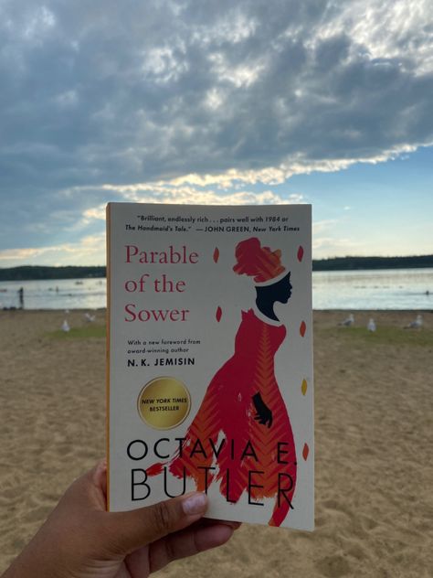 Parable of the Sower cover is held in photo with beach and water in the background. Clear summer skies Octavia E Butler, Parable Of The Sower, Dystopian Novels, Toni Morrison, Lifetime Achievement Award, Handmaid's Tale, Book Recs, John Green, New York Times