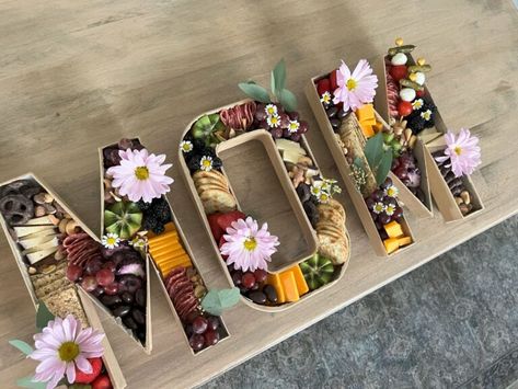 Cardboard letters spelling out MOM and stuffed with snacks and decorated with flowers. Fruit Tray Ideas For Mothers Day, Mother’s Day Hosting Ideas, Mother’s Day Charcuterie Brunch, Mother’s Day Ideas Table, Mother’s Day Bbq Decor, Mother’s Day Lunch Appetizer, Mother’s Day Fruit Tray Ideas, Mom Charcuterie Board, Mother’s Day Lunch Picnic