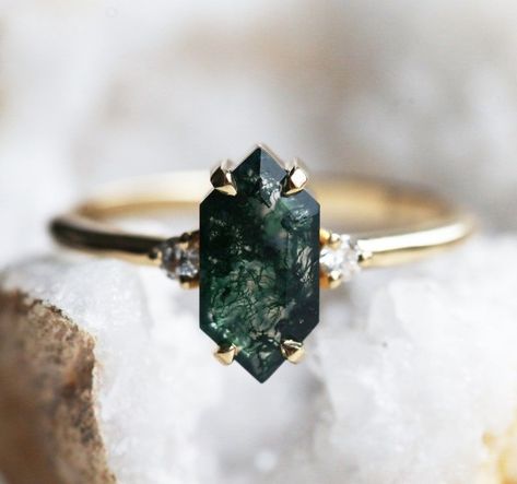 This striking hexagon-cut moss agate engagement ring features vibrant green mossy hues and sparkling round diamond accents.  A unique three-stone design, it's available in various precious metal options and custom gemstone choices.  

Discover matching bracelets, necklaces, and bands to complete the set. Noir Romance, Tiktok Wedding, Ring Hexagon, Green Gemstone Ring, Bling Ring, Cute Engagement Rings, Future Engagement Rings, Dark Wedding, Agate Engagement Ring