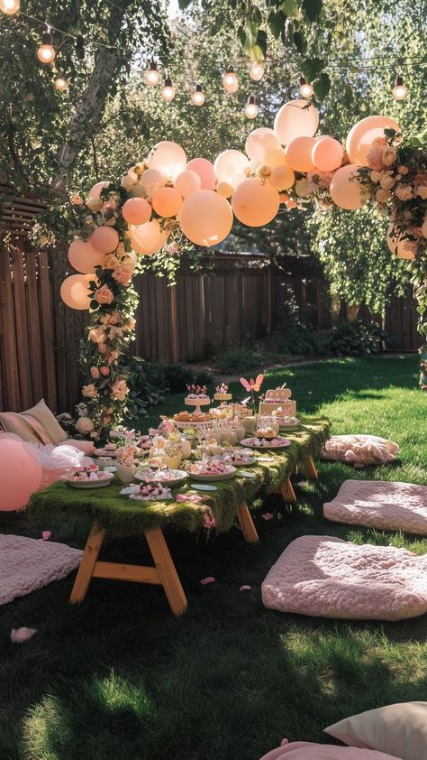 Outdoor fairy garden birthday party setup with a moss-covered table, soft pink cushions, whimsical balloon arch, string lights, and delicate floral decorations, creating a magical and enchanting atmosphere. Leaf Birthday Theme, Outdoor Fairy Birthday Party Ideas, Fairy Garden Gender Reveal, Secret Garden Decorations Party, Rose Garden Birthday Theme, Girls Garden Party Birthday, 1st Birthday Garden Theme, Baby Girls Birthday Theme Ideas, Whimsical Garden Theme Party