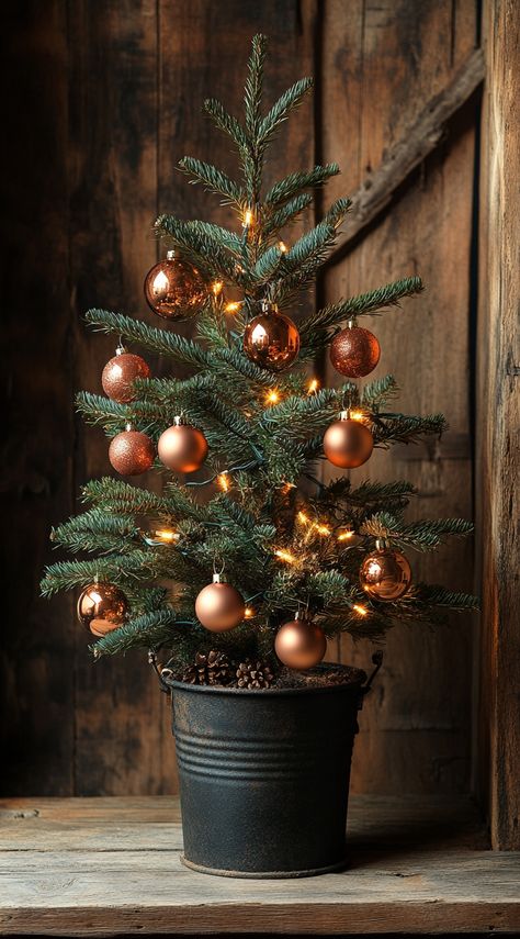 Small Christmas tree in an iron bucket, adorned with copper ornaments and lights, against a rustic wooden backdrop. Christmas Brown Aesthetic, Rustic Woodland Christmas Tree, Earthy Christmas Tree, Forest Christmas Decor, Boho Christmas Decor Ideas, Woodland Christmas Tree, Wooden Backdrop, Chic Christmas Decor, Boho Christmas Decor