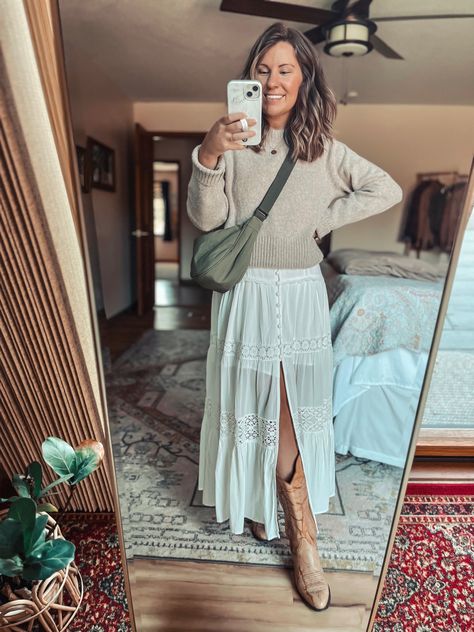 Woman wearing a Long white dress with a sweater over it and cowboy boots. Carrying a green crescent bag. Boho Boots And Dress, Long Dresses With Cowgirl Boots, Boho Cowboy Boots Outfit, Maxi Skirt With Cowboy Boots, Tan Cowgirl Boots Outfit, Prairie Skirt Outfit, Cowgirl Boots Outfit Fall, Tan Cowboy Boots Outfit, Spring Boots Outfit