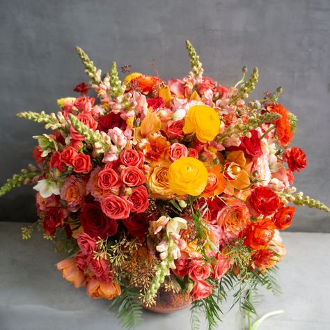 This dual-toned arrangement is ablaze with a gradient of colors, featuring a lush and vibrant bouquet a-top an exquisite underwater scene. The fiery orange tones of spray roses, free spirit roses, shimmer roses, and red roses are complemented by the warmth of orange tulips and cymbidium orchids. The addition of red butterfly ranunculus, yellow ranunculus, and snapdragons adds an extra burst of color, while the seeded eucalyptus and winter roses provide a perfect green contrast. The stunning vase Tall Bouquet Centerpiece, Orange Floral Bouquet, Red Orange Yellow Flower Arrangement, Artichoke Floral Arrangement, Yellow And Red Flower Arrangements, Sunset Color Flower Arrangements, Red Orange Yellow Flowers, Red Roses Flower Arrangement, Ranaculus Wedding Bouquet