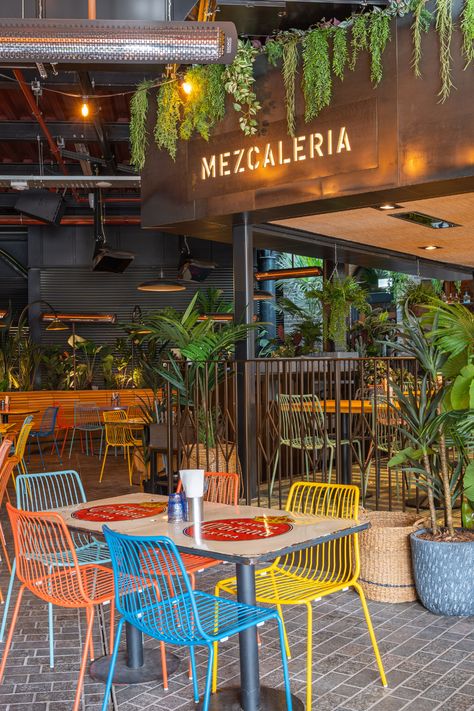 bright coloured seating area with square glossy wooden table, floral background with a mixture of different plants creating a beautiful atmosphere. big open plan restaurant and bar area with black walls and stone floor. Mexican Restaurant Outdoor Patio, High End Mexican Restaurant, Mexican Taqueria Design, Aesthetic Mexican Restaurant, Mexican Cafe Design, Modern Mexican Restaurant Design, Mexican Restaurant Exterior, Cafe Exterior Design Coffee Shop, Family Restaurant Design