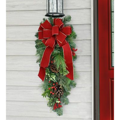 This beautiful pine door swag can be used on a door, an outside light fixture, mantel decoration, stair railings or windows to enhance your holiday season. Get more than one! Created with artificial pine, pine cones, berries and a handmade red velvet bow with gold edge. The Holiday Aisle® | The Holiday Aisle® Faux Pine Teardrop Swag 36.0 H x 5.0 W x 10.0 D in green / red | 30" H X 9" D X 10" L | Wayfair Christmas Swag Outdoor Light, Porch Light Christmas Swag, Red And Green Outdoor Christmas Decor, Lantern Swags Christmas, Christmas Swags For Outdoor Lights, Swag Door Wreath, Christmas Swag For Outdoor Lights, Indoor Christmas Door Decorations, Front Door Decor For Christmas