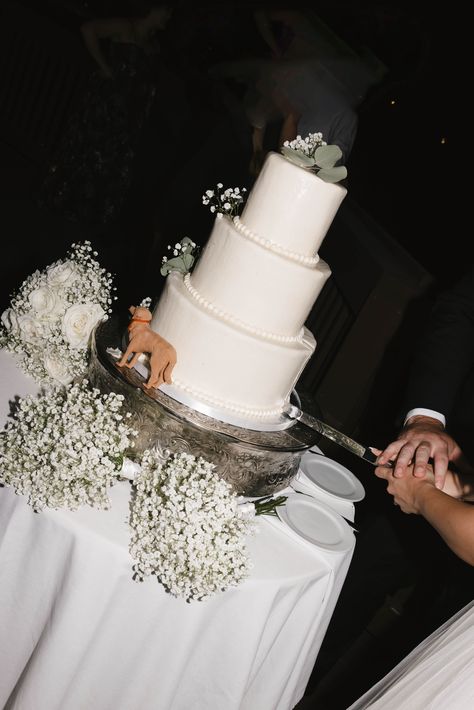 A four-tier white buttercream wedding cake sits on a silver stand, adorned with delicate baby's breath flowers and eucalyptus leaves for a romantic, minimalist design. A small dog figurine adds a playful, personalized touch to the cake's base. The couple's hands, intertwined, are captured mid-slice as they cut the cake together. The scene is softly lit, with white floral bouquets placed around the table, enhancing the elegant, timeless wedding atmosphere.